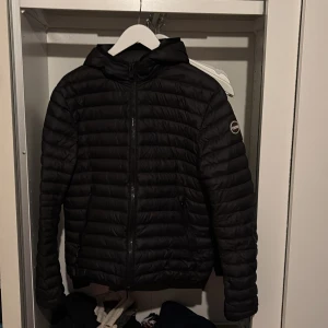 Svart Colmar pufferjacka med huva - Snygg svart pufferjacka från Colmar med huva och dragkedja framtill. Jackan har ett stilrent, quiltat mönster och klassisk logga på ärmen. Perfekt för kyliga dagar och enkel att matcha med allt. Lätt och smidig att bära.