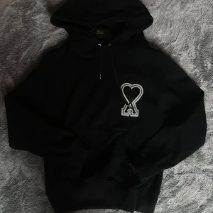 Svart hoodie från Ami Paris med hjärtdetalj - Svart hoodie från Ami Paris med stor broderad logga i form av ett hjärta och bokstaven A på bröstet. Tröjan har huva med snörning och klassisk känguruficka. Perfekt för dig som gillar stilrena streetwear-plagg med unik detalj. Storleken är lite skum!