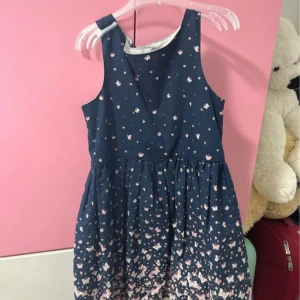 Blommig marinblå klänning Young Dimension - Söt marinblå klänning från Young Dimension med ljusrosa och vita blommor och fjärilar. Klänningen är ärmlös, har rund hals och knappar i ryggen. Kjolen är lätt utställd och materialet känns mjukt och bekvämt, troligen bomull. Perfekt för dig som gillar gulliga detaljer!