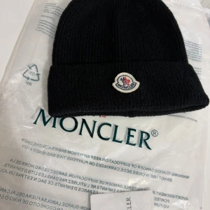 Svart Moncler mössa  - Svart ribbstickad mössa från Moncler med klassisk uppvikt kant och broderad logotyp framtill. Tillverkad i mjuk ull som håller dig varm under kyliga dagar. Perfekt accessoar för en stilren och sportig look.
