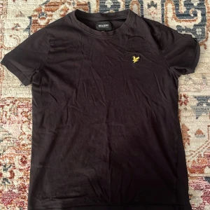 Svart t-shirt från Lyle & Scott Junior - Svart t-shirt från Lyle & Scott Junior med klassisk rund hals och korta ärmar. På bröstet sitter den ikoniska gula örnloggan som broderad detalj. T-shirten är gjord i mjuk bomull och har en clean, enkel look som funkar till allt.