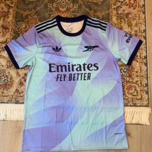 Arsenal x Adidas blå/lila fotbollströja M - Snygg Arsenal fotbollströja från Adidas i slim fit, storlek M. Tröjan har korta ärmar, mörkblå krage och detaljer, samt ett unikt blått och lila mönster. Materialet är lätt polyester med Aeroready-teknologi. Tryck med Emirates och Arsenal-logga på bröstet.