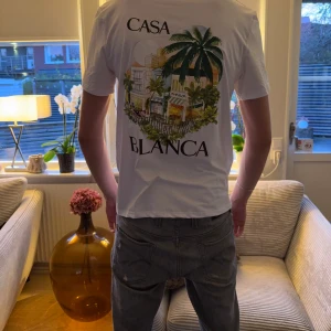 Vit Casa Blanca t-shirt med tryck - Snygg vit t-shirt från Casa Blanca med färgstarkt tryck av palmer och byggnader på ryggen samt mindre tryck på bröstet. Klassisk passform och korta ärmar, perfekt för dig som gillar streetwear med lite extra vibe.