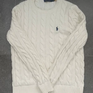 Vit kabelstickad tröja Polo Ralph Lauren - Kabelstickad vit tröja från Polo Ralph Lauren med klassisk rund hals och broderad marinblå logga på bröstet. Tröjan är i mjuk bomull och har ribbade muddar vid ärmslut och nederkant. Perfekt för lager-på-lager under hösten och vintern. Bara testad!