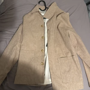 Beige kavaj med knappar från Jack and jones - Snygg beige kavaj med klassisk krage och tre stora knappar framtill. Jackan har två rejäla fickor och är tillverkad i ett linne-liknande material som ger en chill och stilren vibe. Perfekt för dig som gillar enkel och clean look.