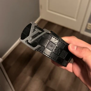 Louis Vuitton rutigt svart bälte - Snyggt svart bälte från Louis Vuitton med klassiskt rutigt mönster och stor LV-logga i silver på spännet. Bältet är gjort i exklusivt skinn och har en bred design som sticker ut. Perfekt accessoar för att lyfta din outfit.