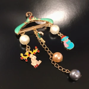 Juligt broschyr med pärlor och berlocker - Unik broschyr med en grön galge som hänge, dekorerat med pärlor och juliga berlocker som en snögubbe, ren och polkagris. Kedjan är guldfärgad och har pärlor i vitt, brunt och grått. Perfekt för dig som vill sticka ut under julen!