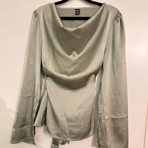 Ljusgrå sidenblus från Shein - Säljer en ljusgrå blus från Shein i glansigt sidenmaterial. Blusen har långärmade ärmar med knappar vid manschetterna, snygg draperad halsringning och ett knytband i midjan som ger en fin siluett. Perfekt för dig som gillar stilrena och eleganta toppar.