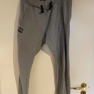 Grå mjukisbyxor från MM Sports - Snygga grå mjukisbyxor från MM Sports i storlek S. Byxorna har ribbad midja med svart snörning, två sidofickor fram och två bakfickor. Materialet är mjukt och stretchigt, perfekt för chill eller träning. Diskret logga på både fram- och baksida.
