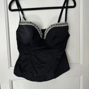 Svart korsett med spetskant - Snygg svart korsett med justerbara axelband och dekorativ vit spetskant vid kuporna i storlek 85C. Toppen har formade kupor och hakknäppning bak för en riktigt cool siluett. Materialet är glansigt och känns lyxigt mot huden. Perfekt för att lyfta din outfit till nästa nivå.