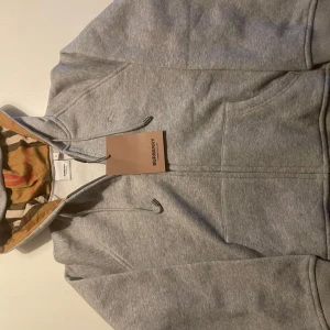 Grå hoodie från Burberry med dragkedja - Snygg grå hoodie från Burberry med klassiskt rutigt foder i huvan och dragkedja framtill. Tröjan har snörning i huvan, känguruficka och är tillverkad i mjuk bomull. Perfekt för dig som vill ha en stilren och lyxig streetwear-look.