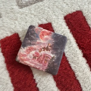 Söt sminkspegel med sagoslottmotiv - En kvadratisk sminkspegel med ett drömmigt motiv av ett slott, rosor och en röd måne på framsidan. Spegeln är i silverfärgad metall och har två speglar inuti, perfekt att ha i väskan. Fantasifull och unik design för dig som älskar sagokänsla.