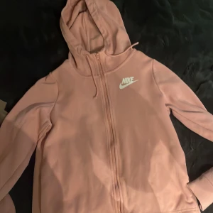 Rosa hoodie från Nike med dragkedja - Säljer en snygg rosa hoodie från Nike i storlek S. Tröjan har huva med snörning, hel dragkedja framtill och Nike-logga i vitt på bröstet. Perfekt för chill eller träning, och tillverkad i mjukt material för extra komfort.