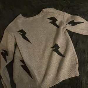 Grå stickad tröja med svarta blixtar - Grå stickad tröja med coola svarta blixtmotiv över hela tröjan. Små svarta hål på baksidan, storlek S 