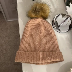  ribbad mössa med pälsboll från ONLY - Superfin beige ribbstickad mössa från ONLY med stor fluffig pälsboll i gult och brunt upptill. Mössan har uppvikt kant och är gjord i mjukt material som håller dig varm och snygg under kalla dagar. Mått: Bredd 21 längd 25 cm 
