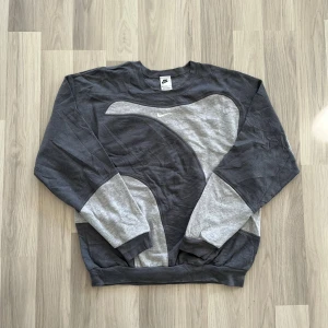 Grå sweatshirt från Nike med mönster - Säljer en grå och ljusgrå sweatshirt från Nike i storlek M. Tröjan har ett stort, böljande mönster framtill och broderad Nike-logga på bröstet. Rund hals och långa ärmar med muddar. Perfekt för dig som gillar sportig och avslappnad stil. Mått; bredd: 50cm, längd: 66cm