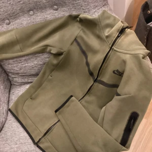 Olivgrön Nike vindjacka med huva - Snygg olivgrön vindjacka från Nike med huva och svarta detaljer. Jackan har hel dragkedja framtill, svarta linjer över bröstet och muddar i svart. Perfekt för dig som gillar sportig stil och vill ha något som sticker ut.