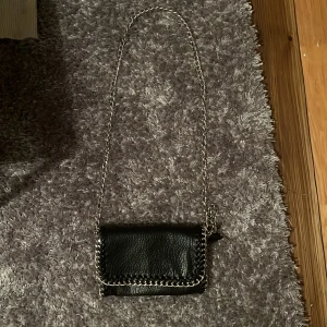 Svart axelväska med kedja - Snygg svart axelväska i skinn med silverfärgad kedja som axelrem. Väskan har en stilren design med kedjedetaljer längs kanten och ett lock som stängs framtill. Perfekt storlek för det viktigaste och ger en cool vibe till din outfit. Nu pris är 500kr.