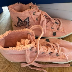 Odd Molly rosa butterfly high sneakers stl 39 - Säljer ett par höga sneakers från Odd Molly i färgen milky pink. Skorna har fransiga kanter, snörning och ett broderat fjärilsmärke på sidan. De är tillverkade i textil med platt sula och metallöglor för snörningen. Perfekt för dig som gillar en unik och cool stil. Liten avfärgning på gummisulan, annars aldrig använda