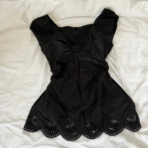 Svart blus med broderad kant Stockh LM - Supersöt svart blus från Stockh LM med broderad och utskuren kant nertill. Blusen har korta ärmar, markerad midja och en snygg rynkning vid bysten. 