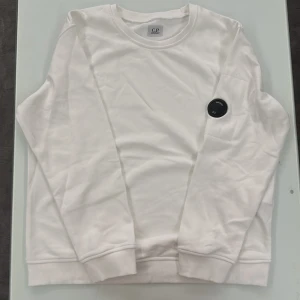 Vit C.P. Company sweatshirt - Vit C.P. Company sweatshirt, lång ärm och ikonisk svart linsdetalj på ärmen. Tröjan har en diskret svart dragkedja på ena armen, Perfekt för vintern ❄️❄️