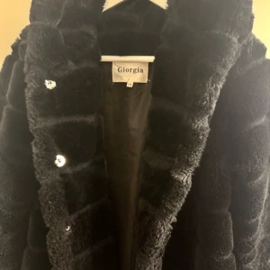 Superfint skick. Svart pälsjacka m stor luva från Giorgia - Nyskick! Säljer en svart otroligt mjuk pälsjacka från Giorgia i storlek S. Slutar precis nedanför rumpan. (På mig iaf) Jackan har fluffig, mjuk yta med markerade paneler och blanka tryckknappar framtill. När jag köpte den så bytte jag ut alla knappar plus la till en högst uppså man kan stänga för mer om man vill. Likaså längst ner.. la till en knapp om man vill stänga för mer vid grenen om man vill.  Insidan är slät och fodrad, perfekt för kalla dagar. Jackan har lång ärm och en oversized look.
