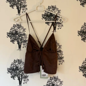 Brun omlott linnetopp med snörning - Supersnygg brun omlott linnetopp från H&M med tunna justerbara axelband och snörning framtill. Toppen har en croppad passform och är tillverkad i glansig polyester som ger en lyxig känsla. Perfekt att styla med jeans eller kjol för en trendig look.