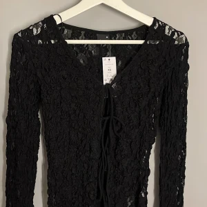 Svart spetsblus med snörning XS - Svart blus i genombruten spets med lång ärm och v-ringad hals. Snygg snörning framtill som ger en cool detalj. Perfekt för dig som gillar att sticka ut med en unik topp. Spetsen har ett blommigt mönster och blusen är figurnära.