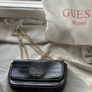 Svart axelväska Guess med kedja - Snygg svart axelväska från Guess med croco-mönstrad yta och guldfärgad kedja som axelrem. Väskan har ett stilrent Guess-emblem i guld på framsidan och beige insida. Perfekt för dig som gillar trendiga detaljer och lyxig känsla.