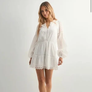 Vit kort klänning med spetsdetaljer - Aldrig använd och prislappen kvar. Superfin vit kort klänning med broderade spetsdetaljer och v-ringad hals. Klänningen har volangkant nertill, ballongärmar och elastisk midja som ger en snygg siluett. Perfekt för sommarens alla tillfällen, riktigt fräsch och trendig look. Nypris 599