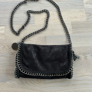 Svart axelväska från Stella McCartney - Cool svart axelväska med kedjerem och snygga silverdetaljer från Stella McCartney. Väskan har ett glansigt yttre i syntetmaterial, dekorativa kedjor runt kanterna och magnetlås. Insidan har ett fack med dragkedja för småsaker.