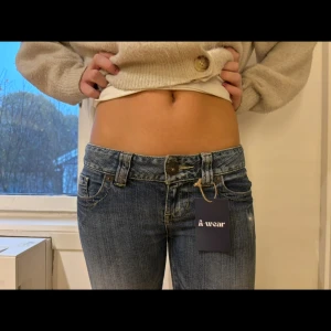 Blå bootcut jeans från Guess - Snygga blå jeans från Guess med låg midja och bootcut passform. Jeansen har klassisk femficksdesign, slitningar och broderade detaljer på bakfickorna. Materialet är jeans med lätt tvättad look och knappstängning framtill.Innerbensmått: 80 cm Ytterbensmått: 90 cm  Midjemått tvärs över: 36 cm  Lårmått: 18 cm  Grenmått: 17 cm (Nr 34)