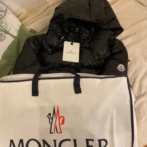 Svart Moncler Maya dunjacka med huva - Svart Moncler Maya dunjacka med glansig finish, huva och dragkedja framtill. Jackan har Moncler-logga på ärmen, två sidofickor med dragkedja och coolt serietryck på insidan. Perfekt för dig som vill ha en snygg och varm jacka med street vibe.