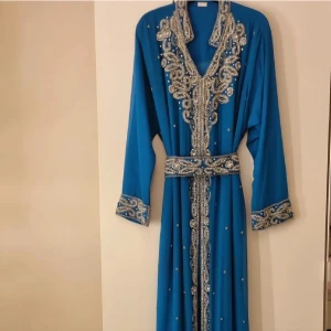 Blå långklänning med silverbroderi - Säljer en blå långklänning med fantastiska silverbroderier och pärldetaljer längs krage, ärmslut och hela framsidan. Klänningen har v-ringning, lång ärm och ett matchande bälte i midjan. Perfekt för dig som vill sticka ut med en elegant och unik look.