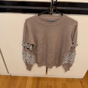 Beige stickad tröja med spetsdetaljer - Snygg beige stickad tröja från Laetitia Mem med unika spetsdetaljer och pärlor på ärmarna. Tröjan har rund hals och långärmade ärmar med ribbade muddar. 