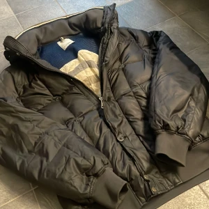 J.Lindberg Dunjacka - Vintern kommer och du behöver en riktigt varm och fet vinterjacka, här har du ditt svar❄️|| En svart dunjacka med hög krage👏🏻👏🏻|| Storlek: L, men sitter mer som M (Modellen 182, 70kg) || Skick; 8/10 (bild 5 defekt), Knapp saknas på framsida men finns extra knappar i jackan || Pris: 799kr