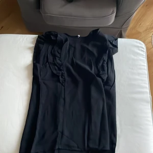 Svart volangklänning med kort ärm - Svart klänning med volangdetaljer längs axlarna och ärmarna. Modellen är ärmlös med breda volangpartier som ger en cool och trendig look. Klänningen har rund halsringning och knapp i nacken. Tunt, lätt material som faller snyggt.