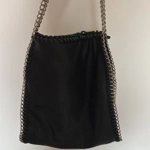 Svart axelväska med kedjedetaljer Gina Tricot - Cool svart axelväska från Gina Tricot i syntetmaterial med silverfärgade kedjor längs kanterna och som axelrem. Väskan har ett stilrent, slätt yttre och dekorativa sömmar runt om. Perfekt storlek för det viktigaste och med innerfack med dragkedja.