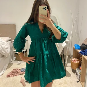 Grön  kortklänning från Zara - Supersöt grön kortklänning från Zara med v-ringning och voluminösa trekvartsärmar. Klänningen har en luftig passform med flera volanger och är tillverkad i bomull. Perfekt för dig som gillar en avslappnad men trendig look.