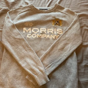 Grå stickad tröja Morris Company XS - Snygg grå stickad tröja från Morris Company i storlek XS. Tröjan har broderad logga och text framtill, rund hals och ribbade muddar vid ärmslut och nederkant. Perfekt för dig som gillar klassisk stil med en modern touch.