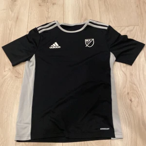 Svart MLS fotbollströja från Adidas - Snygg svart fotbollströja från Adidas med MLS-logga på bröstet och klassiska Adidas-ränder på axlarna. Tröjan har gråa paneler längs sidorna och är tillverkad i lätt, ventilerande Aeroready-material som passar perfekt för träning. Rund hals och korta ärmar.