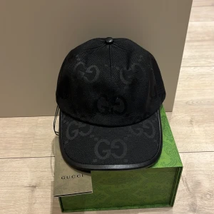 Svart keps från Gucci med GG-mönster - Snygg svart keps från Gucci med diskret GG-logomönster över hela kepsen. Tillverkad i polyester med detaljer i skinn och justerbar rem baktill. Kommer med originalkartong och tagg. Perfekt för dig som vill ha en lyxig streetstil.