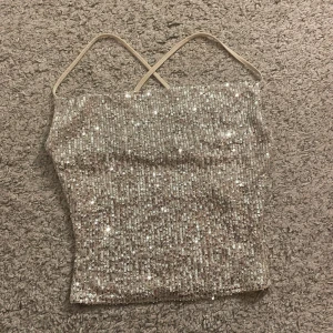 Glittrig topp från NA-KD - Säljer en glittrig och snygg silverfärgad topp från NA-KD med tunna axelband som korsas i ryggen. Toppen är täckt av paljetter och har en croppad passform. 