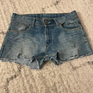 Blå jeansshorts med slitningar - Säljer ett par blå jeansshorts med råa, fransiga kanter och slitna detaljer både fram och bak. Klassisk femficksmodell med knapp och dragkedja. Perfekt för dig som gillar en chill och edgy stil. Passar till sneakers eller boots.
