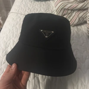 Svart bucket hat från Prada - Säljer en svart bucket hat från Prada med ikonisk triangellogo i metall framtill. Hatten har klassisk form med bred brätte och är tillverkad i ett glansigt syntetmaterial med slät finish. Insidan är fodrad för extra komfort och har en diskret Prada-etikett.