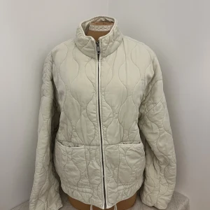 Quiltad beige pufferjacka med dragkedja - Quiltad pufferjacka i beige med snyggt vågigt mönster. Jackan har hög krage, två stora fickor framtill och dragkedja hela vägen. Materialet känns mjukt och jackan har en loose passform som ger chill vibe. Perfekt för dig som gillar stilren och enkel design.