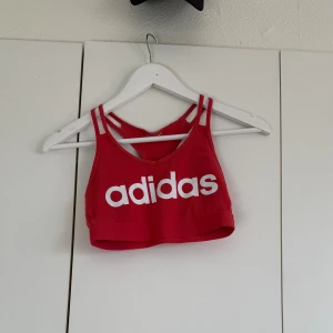 Röd linnetopp från Adidas - Säljer en röd linnetopp från Adidas med vit logga framtill. Toppen har dubbla smala axelband och en racerback-design som ger en sportig vibe. Perfekt för träning eller chill dagar. Materialet är mjukt och stretchigt för skön passform.