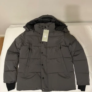 Grå pufferjacka från Canada Goose - Snygg grå pufferjacka från Canada Goose med huva och svart logotyp på ärmen. Jackan har svarta knappar, dragkedja och ribbade muddar vid ärmslut. Fodret är svart och jackan är tillverkad i polyester med dunfyllning. Perfekt för kalla vinterdagar.