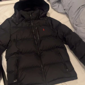 Svart pufferjacka från Polo Ralph Lauren - Svart pufferjacka från Polo Ralph Lauren med huva, flera dragkedjeförsedda fickor och klassisk röd logga på bröstet. Jackan är tjockt vadderad och har en modern, loose passform som är perfekt för kalla dagar. Snygg och stilren design med hög krage och detaljerade sömmar. OBS!! Storleken är XL i barnstorlek och det motsvarar Vanlig small inget fel på den alls helt ny 