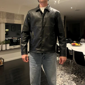 Svart skinnjacka från Zara - Snygg svart skinnjacka från Zara med klassisk krage och raka linjer. Jackan har knappar framtill och två sidofickor. Varm nog för vintern.